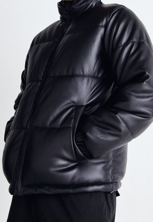 Homme portant une veste en cuir matelassée noire avec une fermeture éclair à l'avant et les mains dans les poches sur un fond clair uni.