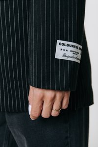 Μαύρο σακάκι με ριγέ σχέδιο και ετικέτα που γράφει "COLOURFUL REBEL" στη μανσέτα. Διαθέτει δομημένο σχέδιο και απαλή υφή υφάσματος.