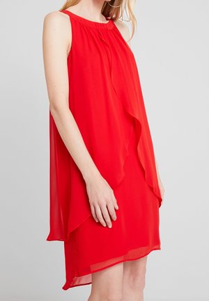 Femme portant une robe sans manches rouge vif à couches avec un tissu fluide drapé à l'avant, sur un fond gris clair uni.