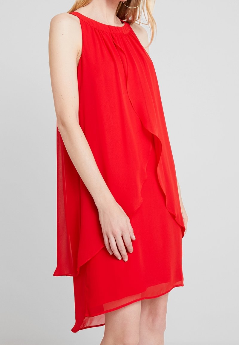 Femme portant une robe sans manches rouge vif à couches avec un tissu fluide drapé à l'avant, sur un fond gris clair uni.