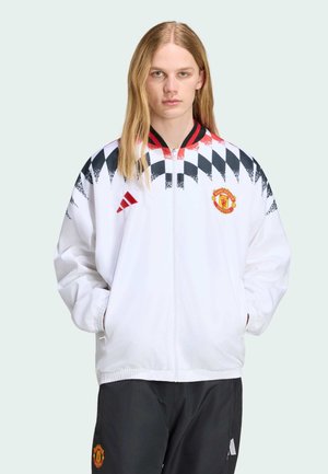 Person with long blonde hair wears white zip-up jacket with black and red chevron pattern and Manchester United logo, paired with black pants.