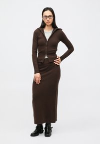 Smeđi pleteni kardigan sa zatvaračem uparен s odgovarajućom dugom suknjom. Nošeno preko svijetlosive majice. Model stoji u crnim gležnjačama.