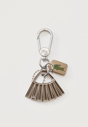 Porte-clés en métal argenté avec breloque en forme d'éventail, fermoir rond et petite étiquette beige ornée d'un logo de crocodile vert sur un fond uni.