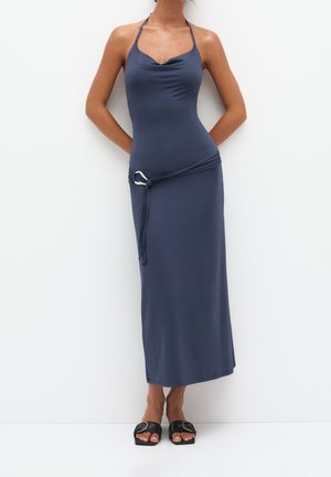 Femme portant une robe maxi sans manches bleu marine à col montant avec une ceinture et des sandales plates noires à bout ouvert, debout sur un fond blanc.