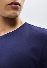 Κοντινό πλάνο του κάτω μέρους του προσώπου και του λαιμού ενός ατόμου που φοράει μπλε σκούρο μπλουζάκι με στρογγυλή λαιμόκοψη, πάνω σε απλό φωτεινό φόντο.