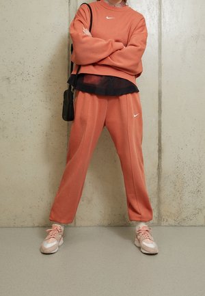Coral sweatshirt with white Nike logo, layered over a dark mesh top. Matching coral sweatpants with side seams. Pink athletic shoes.