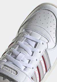 White leather sneaker featuring perforated details, white laces, with red and grey stripe accents on the side and a textured outsole.