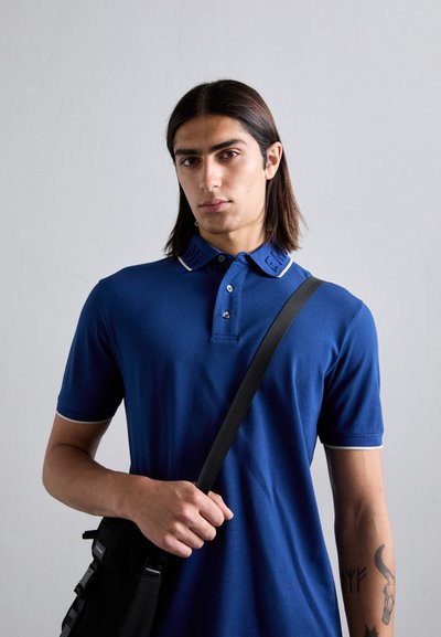 Tânăr cu păr lung și închis la culoare, purtând un tricou polo albastru și o geantă neagră de umăr, stând în fața unei pereți de culoare gri deschis.