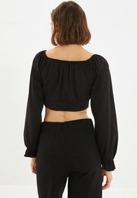 Schwarzes, kurzes Oberteil mit langen, gepufften Ärmeln und elastischem Ausschnitt, kombiniert mit hochtaillierten schwarzen Hosen, die beide aus einem glatten Stoff bestehen.