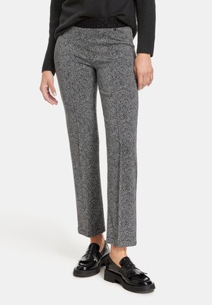 Femme portant un pantalon droit en chevrons gris, un pull noir et des mocassins en cuir verni noir, debout devant un fond blanc.