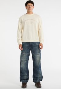 Cremefarbener Strickpullover mit gerippten Bündchen und dem Schriftzug "Guess Originals". Getragen mit dunkelblauen Cargo-Jeans mit mehreren Taschen und lockerer Passform.