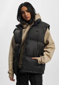 Schwarze Steppweste mit Reißverschluss und Seitentaschen, über einem beigen Kapuzensweatshirt getragen. Glattes Material und gestepptes Design.