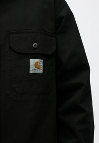 Μαύρη τσέπη στο μπουφάν Carhartt με καπάκι που κλείνει με κουμπί και μικρό διάτρητο λογότυπο σε καφέ και λευκό.