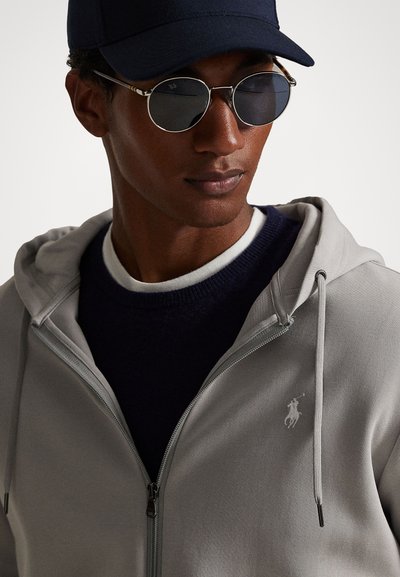 Corta-vento cinza com fecho éclair e capuz com cordão, logótipo branco de polo, usado sobre um suéter crewneck azul-marinho. Óculos de sol circulares escuros e um boné azul-marinho completam o visual.