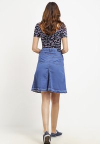 Falda de mezclilla en línea A con dobladillo deshilachado, de color azul y de longitud media. Presenta bolsillos traseros y una textura suave, combinada con zapatillas casuales.