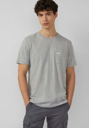 Jeune homme portant un t-shirt gris clair à manches courtes et un pantalon cargo gris foncé, debout les mains dans les poches devant un fond uni.