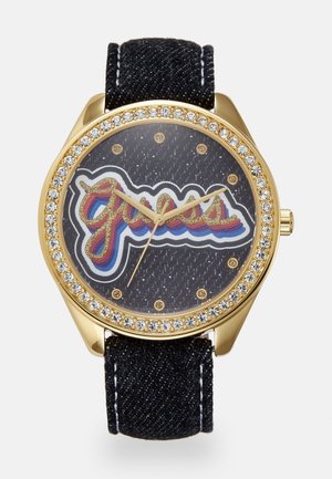 Montre en or avec lunette ornée de cristaux, bracelet en tissu noir, et logo "Guess" multicolore sur un cadran noir texturé avec des index horaires dorés.