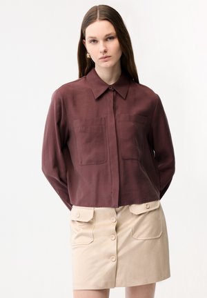 Młoda kobieta o długich brązowych włosach, ubrana w bordową koszulę zapinaną na guziki i beżową zapinaną spódnicę z patkami na kieszeniach, stojąca na jednolitym tle.