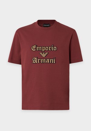 Άνδρας που φοράει κόκκινη μπλούζα Emporio Armani, καφέ παντελόνια φαρδιά και μαύρα παπούτσια, στέκεται με το ένα χέρι στην τσέπη.