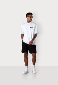 White cotton t-shirt with short sleeves and a graphic logo, paired with black shorts. Worn with white sneakers and dark sunglasses.