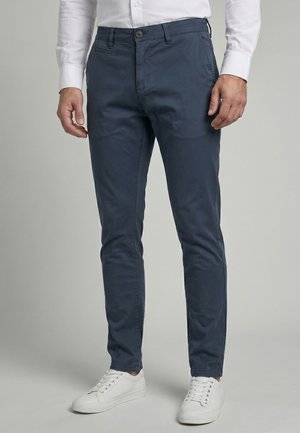 Homme portant un pantalon bleu marine slim, une chemise blanche à manches longues et des baskets blanches, debout devant un fond uni.