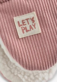 Tessuto rosa a costine con fodera bianca e morbida. Presenta una patch rettangolare tessuta con la scritta "GIOCHIAMO" in caratteri rosa chiaro.