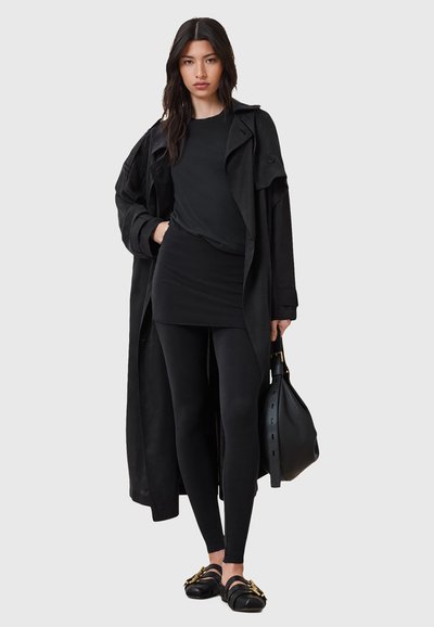 Manteau noir sur un haut noir, petite jupe et leggings, avec des chaussures noires ornées d'accents dorés et un sac texturé.