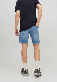 Man standing with one hand in back pocket, wearing rolled-up light blue denim shorts, black t-shirt, white socks, and gray sneakers.