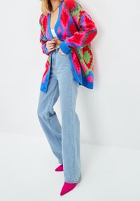 Kleurijke gebreide cardigan met geometrische patronen in rood, groen en blauw. Gecombineerd met lichtblauwe jeans en felroze pumps met een spitse neus.