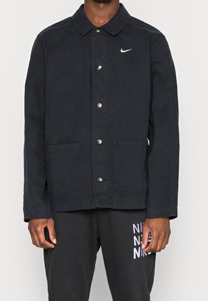 Mann trägt schwarze Jacke mit Knöpfen, drei vorderen Taschen und weißem Nike-Logo auf der Brust, kombiniert mit schwarzen Nike-Sweatpants.