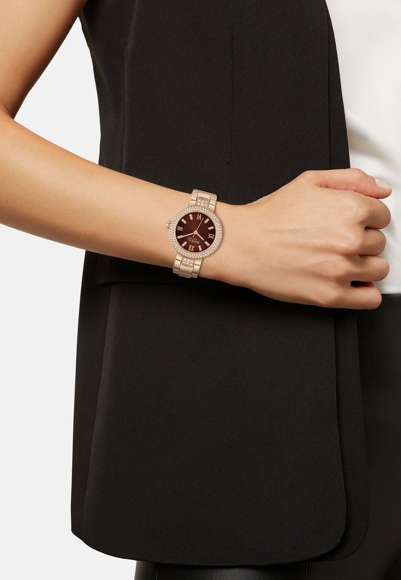 Montre en or rose avec un cadran rond brun, des chiffres romains et des accents en cristal sur la lunette. Bracelet en maillons métalliques avec une texture lisse.