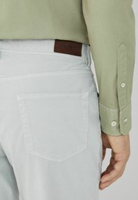 Hellgraue Baumwollhose mit einer Gesäßtasche und einem braunen Lederlabel. Das Hemd ist olivgrün mit geknöpften Manschetten.
