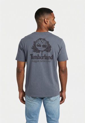 Homme portant un T-shirt Timberland gris foncé avec le logo de la marque et le texte "Rugged Active Gear" au dos, associé à un jean bleu clair.