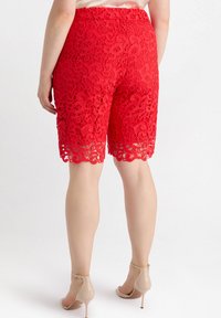 Pantaloni corti rossi in pizzo con un motivo floreale, taglio a metà lunghezza e orlo scallop. Il design aggiunge profondità grazie a dettagli intricati.
