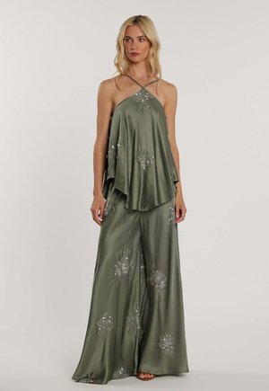 Donna bionda che indossa un top halter in raso verde oliva e pantaloni a gamba larga abbinati con motivi floreali di paillettes su sfondo semplice.