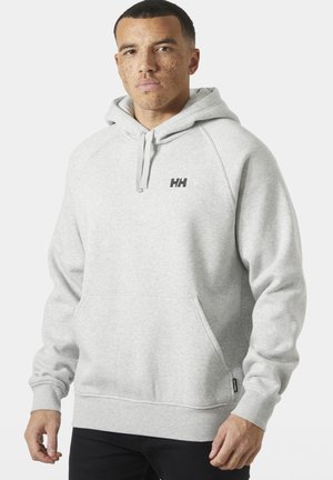 Jonge man met kort haar en sproeten, draagt een lichtgrijze hoodie met het "HH"-logo op de borst en een zwarte broek, staand tegen een witte achtergrond.