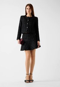 Black tweed jacket with cropped design, round neckline, and large buttons, paired with a matching skirt. The model holds a small burgundy bag.