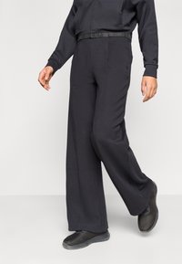 Άτομο που φοράει μαύρο παντελόνι φαρδιάς γραμμής, μαύρο μπουφ άν με φερμουάρ και μαύρα πάνινα παπούτσια χωρίς κορδόνια, περπατώντας μπροστά σε απλό φόντο.