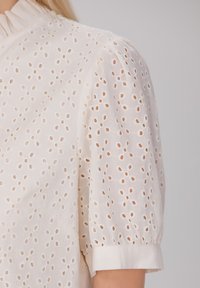 Blouse blanche en broderie anglaise avec un col volanté, des manches courtes bouffantes et un motif délicat de découpes circulaires sur tout le tissu.