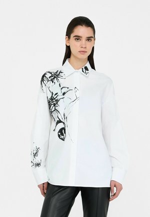 White button-up shirt featuring black botanical embroidery on one side and black collar accents. Long sleeves, relaxed fit, and smooth texture.