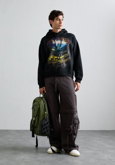 Felpa nera con stampa grafica, pantaloni grigi larghi e uno zaino verde con accenti neri. Sneakers bianche completano l'outfit.