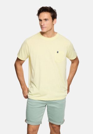 Hombre con camiseta de manga corta amarillo pálido con bolsillo en el pecho y pantalones cortos verdes claros remangados, de pie con las manos en las caderas.