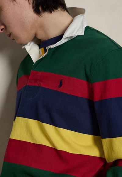 Άτομο που φοράει ρουά μπλούζα με γιακά με οριζόντιες ρίγες σε πράσινο, κόκκινο, ναυτικό και κίτρινο χρώμα, και με μικρό κεντημένο λογότυπο παίκτη πόλο στο στήθος.