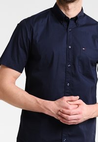 Granatowa koszula z krótkim rękawem na guziki z kołnierzykiem, przednią kieszonką i pięcioma guzikami. Materiał jest gładki z subtelnym połyskiem i bez widocznych wzorów.
