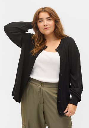 Sort cardigan med lange ærmer og knaplukning, lavet af et blødt materiale. Bæres over en hvid tanktop og olivengrønne bukser.