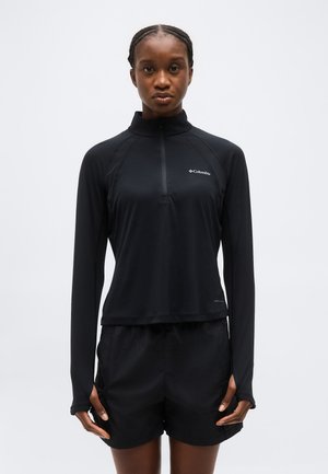 Žena s vrkočmi, oblečená v čiernom športovom tričku s dlhým rukávom značky Columbia a čiernych šortkách, stojaca pred jednoduchým pozadím.