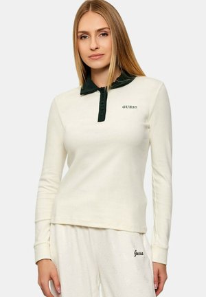 Donna con capelli biondi lisci che indossa un top bianco a maniche lunghe con colletto nero e logo "Guess", abbinato a pantaloni bianchi a trama.