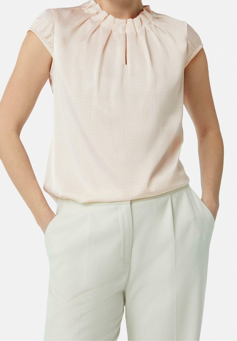Femme portant un chemisier rose pâle à manches courtes avec des plis et une encolure en trou de serrure, rentrée dans un pantalon blanc taille haute, mains dans les poches.