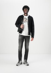 Čierny rebrovaný kardigán cez grafické biele tričko, skombinované s tmavými džínami a viacfarebnými teniskami. Nosiac čiernu crossbody tašku.