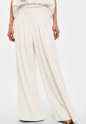 Donna che indossa pantaloni color crema a vita alta e gamba larga con cintura e pieghe, abbinati a un top chiaro, in piedi su sfondo bianco.